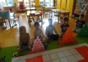 Dzieci siedzą przed ułożonymi z kubeczków kolorowymi wieżami.